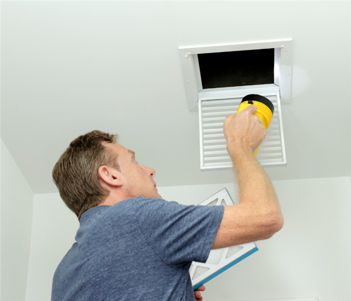 Picture shows a man on a ladder looking inside his ceiling vent to change the furnace filter he is holding. 