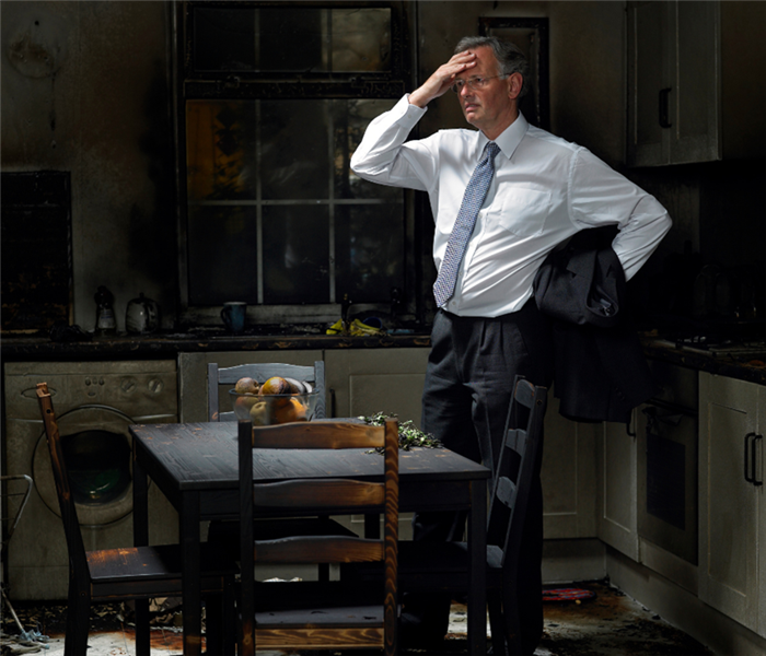 Picture is of a man standing in his burnt down kitchen looking distraught with his hand on his head. 